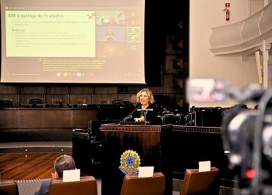 Fotografia de uma mulher em palestra no plenário do TRT-PR, ela fala da tribuna com imagem no telão