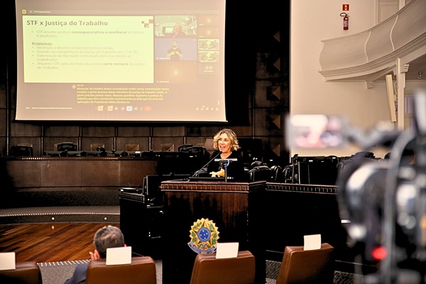 Fotografia de uma mulher em palestra no plenário do TRT-PR, ela fala da tribuna com imagem no telão